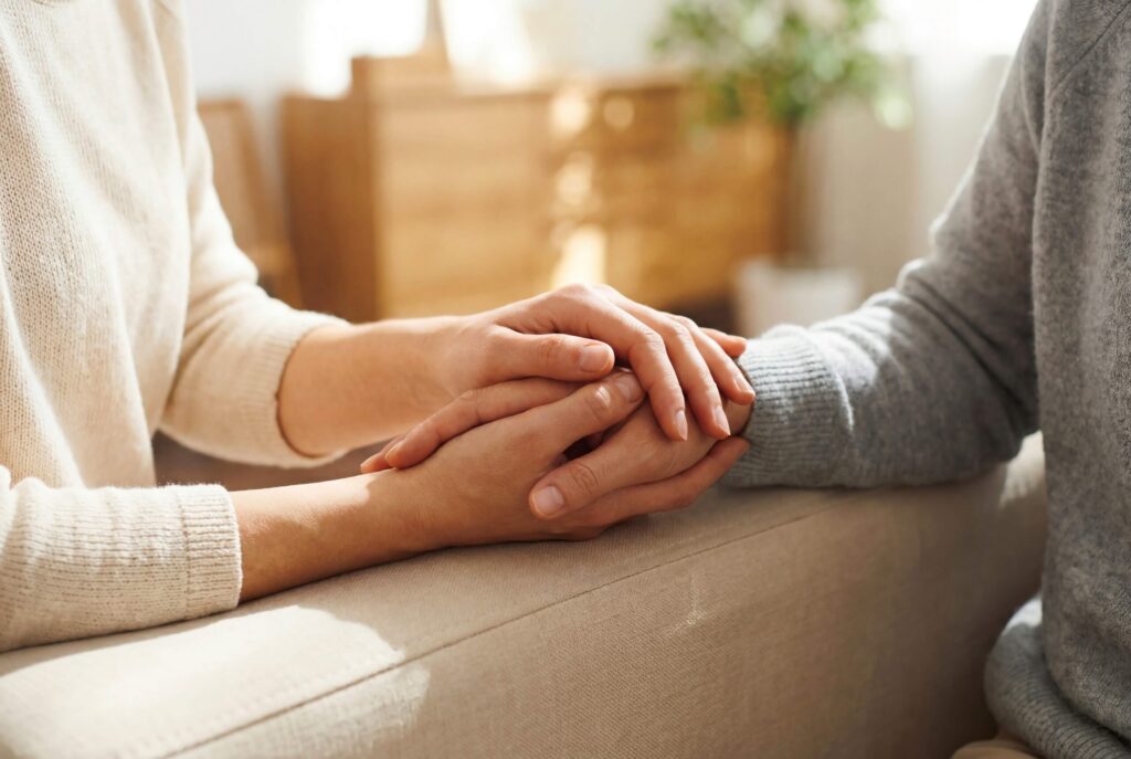 Two people sitting on a couch placing their hands together in a gesture of support and connection.